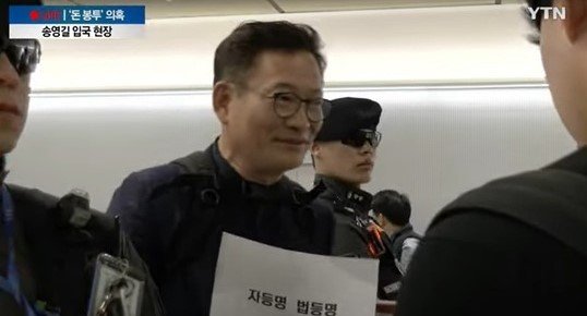 지난 24일 인천공항에 도착한 송영길 전 더불어민주당 대표가 한 여성으로부터 '자등명법등명'이라고 적힌 A4 용지 크기의 종이를 한 장 받았다. (YTN 보도화면 갈무리)
