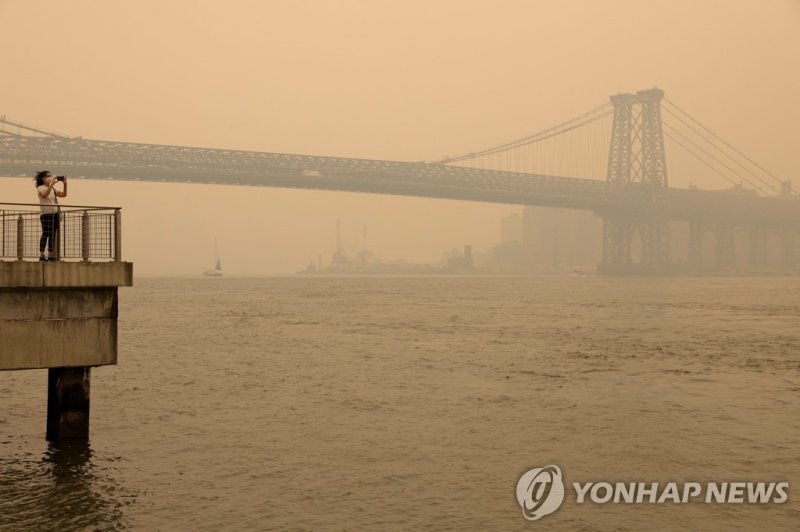 연기로 뿌옇게 뒤덮인 뉴욕 맨해튼 일대 A person wearing a face mask takes a picture of the Manhattan skyline while it's covered in haze and smoke caused by wildfires in Canada, in Brooklyn, New York, United States, June 7, 2023. REUTERS/Amr Alfiky