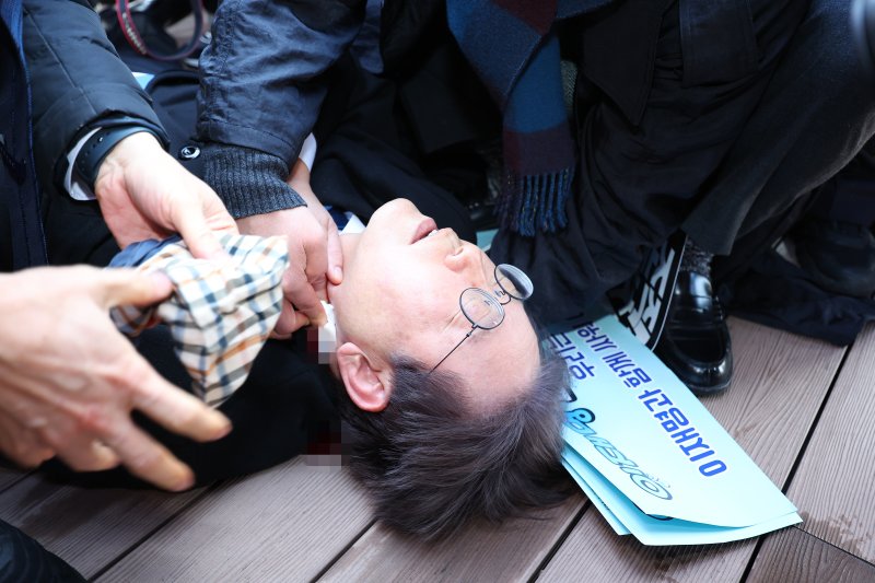 더불어민주당 이재명 대표가 지난 1월 2일 부산 가덕도 신공항 부지를 둘러본 후 기자들과 문답을 진행하던 중 왼쪽 목 부위에 습격을 당해 피를 흘린 채 쓰러져 있다.<div id='ad_body3' class='mbad_bottom' ></div> 사진=연합뉴스