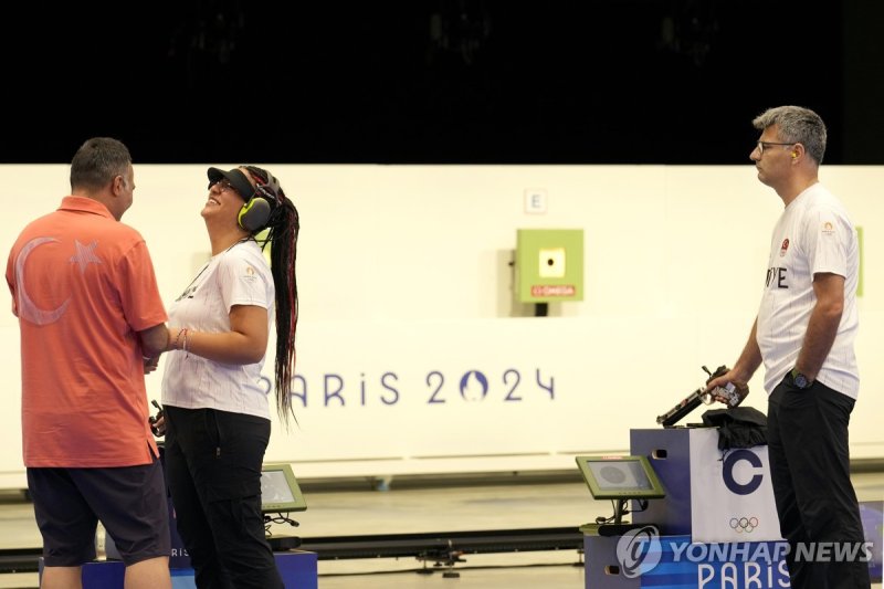 Turkish shooter Yusuf Dikec, right, looks on as Turkey's coach talks to Dikec's teammate Sevval Ilayda Tarhan during a time-out break at the 10m air pistol mixed team final, in Chateauroux, France, at the 2024 Summer Olympics, Tuesday, July 30, 2024. The Turkish team won the silver medal. (AP Photo/