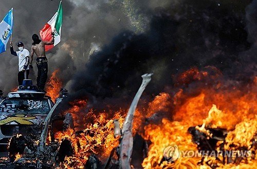 LA에서 차량이 불타고 있는 가운데 깃발을 든 불법이민자 단속 반대 시위대 (출처=연합뉴스)