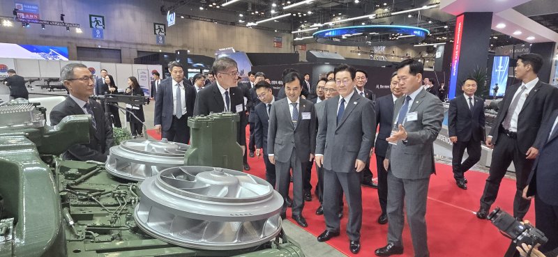 이재명 대통령이 20일 ADEX 2025 전시회에서 SNT그룹 부스를 방문해 K2전차 4차 양산 적용 예정인 국산 파워팩에 대한 설명을 듣고 있다. SNT그룹 제공