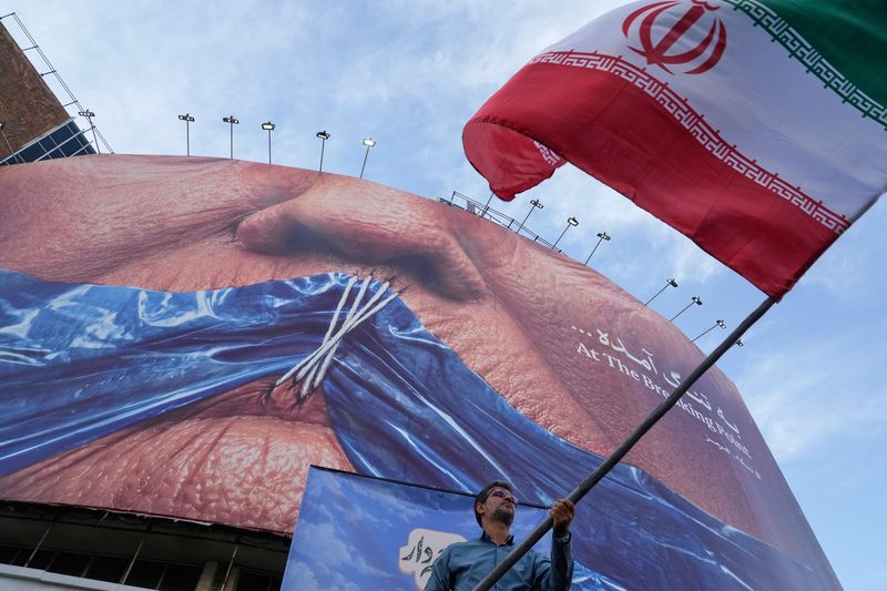 [테헤란=AP/뉴시스] 사진은 지난 6일(현지 시간) 이란 테헤란 도심 광장에서 열린 친정부 집회 참가자가 이란 국기를 흔들고 있는 모습. 이 남성 뒤에는 호르무즈 해협이 도널드 트럼프 미국 대통령의 입을 꿰맨 모습이 담긴 대형 홍보물이 걸려 있다. 2026.05.20.